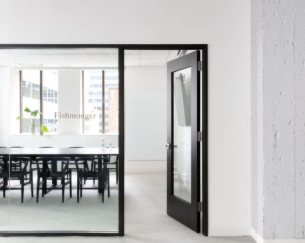 Bright contemporary commercial office interior with a glazed meeting room or dining-style conference area visible through black-framed glass partitions. The space has white walls, pale flooring, exposed textured concrete at the right, black-framed doors, and a long table with multiple black chairs. Large windows provide abundant daylight and views of neighbouring urban buildings.