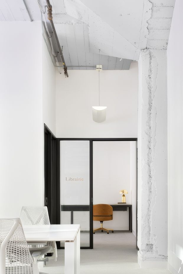 Bright minimalist office interior with white walls and light flooring, exposed unfinished concrete ceiling and services, and a rough concrete structural column. A black-framed glazed partition separates the main workspace from a small private office or study area with a desk, warm brown task chair, and lamp. White mesh office chairs and a white table occupy the foreground, while a simple white pendant light hangs centrally.