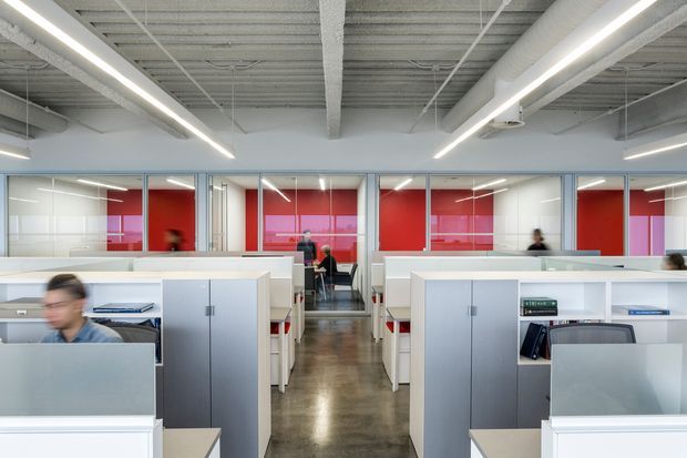 An open-plan contemporary office interior with rows of white workstation partitions and built-in storage, glass-fronted meeting rooms or offices at the rear, and vivid red feature walls. The space has an exposed industrial-style ceiling with visible services, linear suspended lighting, and a polished concrete floor. Several blurred occupants are present.