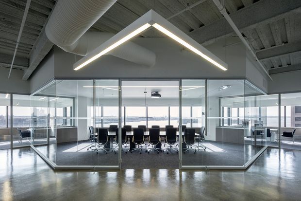 A modern commercial office interior centered on a glass-enclosed boardroom. The room contains a long conference table surrounded by black office chairs, with expansive glazing and a waterfront or urban view beyond. The open office around it has polished concrete flooring, exposed structural ceiling elements, visible HVAC ductwork, and a large geometric linear pendant light. The palette is predominantly cool grey, white, black, and clear glass, with a bright, refined, contemporary workplace atmosphere.