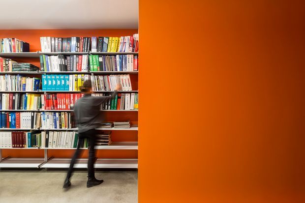 A contemporary library or bookshop interior with floor-to-ceiling open shelving densely filled with books on the left and a large vivid orange wall or partition occupying the right side. A motion-blurred person reaches toward the shelves. The space has a light neutral ceiling and floor, strong orange-and-red accents, and a graphic, minimalist composition.