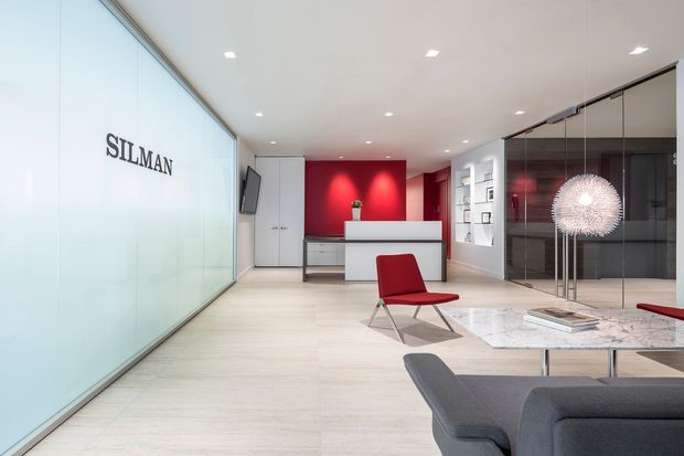 A bright, contemporary commercial reception and waiting area with a frosted glass partition bearing the SILMAN name, pale timber-look flooring, a white reception desk backed by a vivid red feature wall, glass-fronted meeting rooms, and modern lounge seating. A sculptural pendant light and red accent chair add visual interest to the refined open-plan interior.