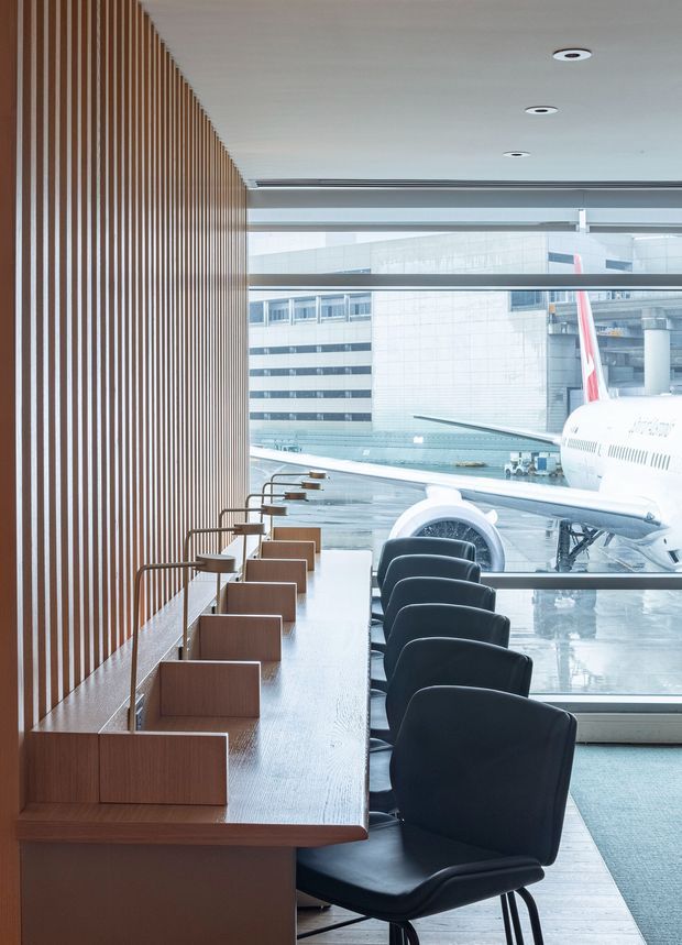 A refined airport lounge or terminal seating area beside expansive glazing overlooking an aircraft and airport apron. A long built-in timber counter with individual study or work stations runs along a vertical timber slat wall, paired with dark upholstered chairs facing the window. The space has a pale ceiling with recessed downlights, polished light flooring, and a minimalist contemporary fit-out.