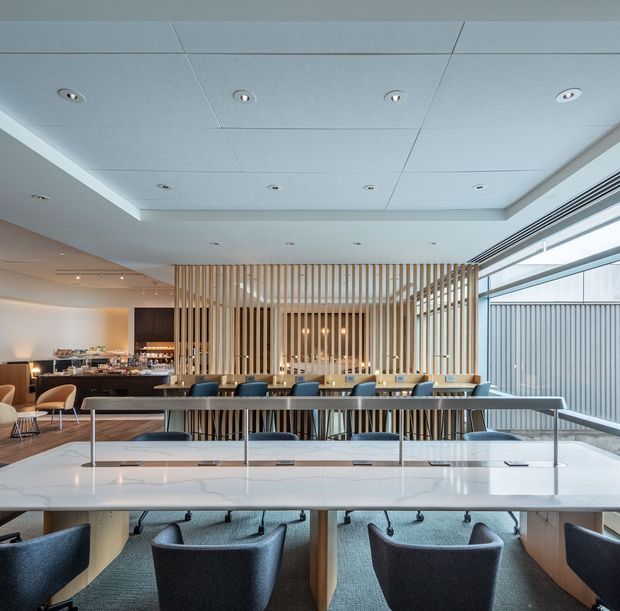Bright contemporary hospitality and dining interior with a long communal table in the foreground, upholstered chairs, and a bar or service counter behind a vertical timber-slat screen. A layered recessed ceiling with many downlights, full-height glazing, pale timber finishes, and neutral upholstery create an airy, refined atmosphere.