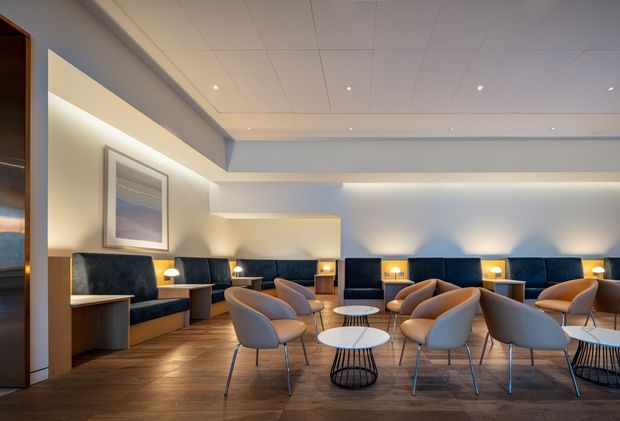 A refined contemporary lounge or waiting area with clusters of upholstered armchairs and low round tables arranged on a dark timber floor. Built-in dark blue banquette seating lines the walls, complemented by warm wood side tables and soft integrated cove lighting. The space has a suspended grid ceiling with recessed spotlights, pale neutral walls, and a framed abstract artwork.