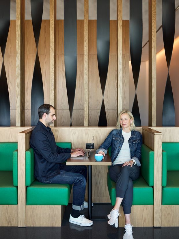 A contemporary commercial-style seating area with two people seated at a small table in green upholstered booth seating. The space features extensive light timber wall panelling and sculptural vertical black geometric panels, with integrated booth joinery and a dark polished floor. A laptop and coffee cups are on the table. The image is tightly framed around the designed interior and may represent a hospitality, workplace, or lounge setting.