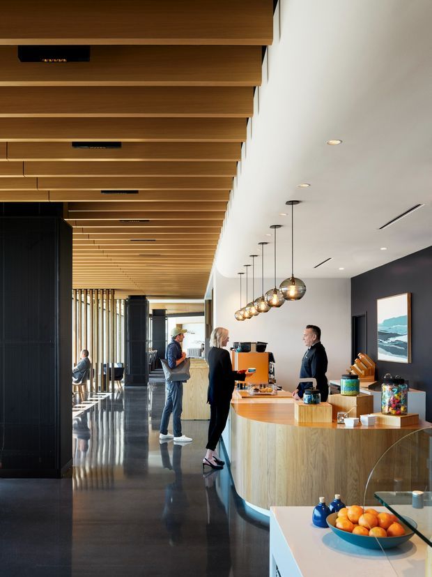 A polished contemporary commercial reception and service counter, likely a café, hospitality venue, or workplace amenity space. The long curved timber-clad counter has a light wood benchtop, coffee equipment, jars, and display items, with pendant globe lights suspended above. A dramatic slatted timber ceiling extends through the space, contrasting with dark wall panels and glossy dark flooring. Floor-to-ceiling glazing and vertical timber screens line the corridor, while people stand at the counter and a framed coastal artwork adds a small visual accent.