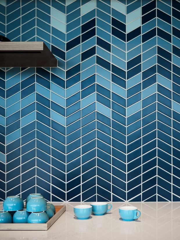 Close-up of a bold blue and pale-blue chevron-patterned tiled wall forming a corner behind a light stone or engineered-stone benchtop. Turquoise ceramic cups and a tray sit on the counter, with a dark timber open shelf projecting from the upper left. The image emphasises geometric tilework and a saturated blue palette.
