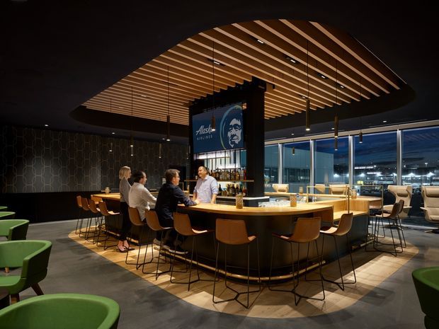 A warmly lit contemporary commercial hospitality interior, likely a bar or restaurant lounge. The focal point is a large curved communal timber bar with integrated counter seating, surrounded by additional lounge chairs and tables. A dramatic oval timber slatted ceiling feature hangs above the bar, with dark ceilings, patterned feature wall cladding, expansive glazing, pendant lighting, and a prominent branded sign. The space has a refined, social evening atmosphere.