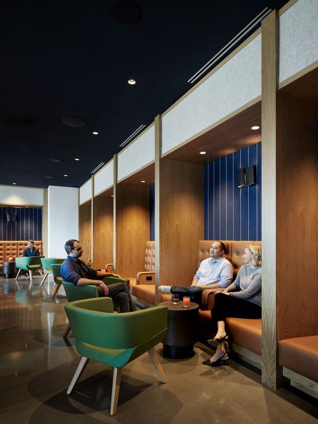 A stylish hospitality or commercial lounge with a series of semi-enclosed timber-lined booth seating areas. The space features deep navy ceilings, warm wood wall and ceiling panels, textured light-coloured upper partitions, blue vertical feature panels, tan upholstered bench seating, and green mid-century-style lounge chairs around small dark tables. Polished flooring, recessed lighting, and occupants relaxing in the booths create a refined, intimate atmosphere.