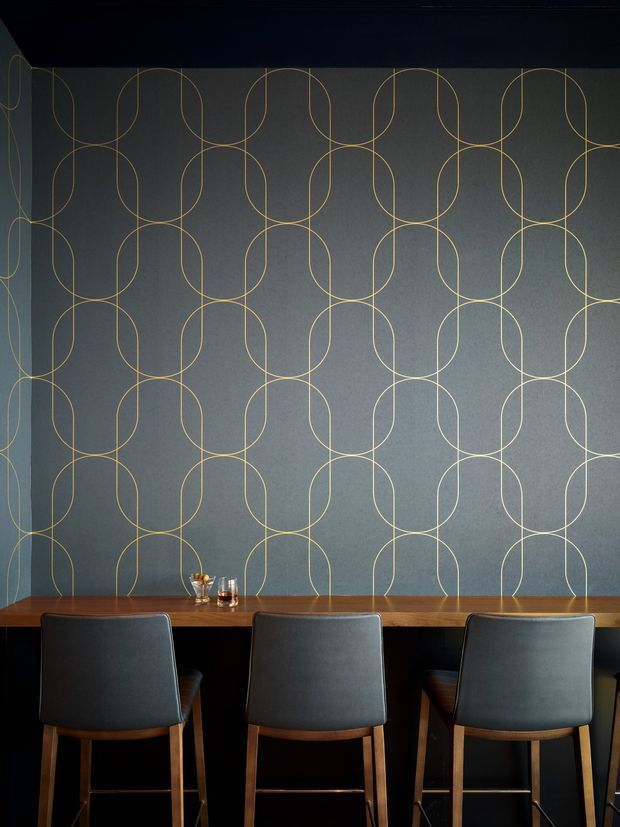 A moody, refined dining or bar-height seating area with three dark upholstered stools arranged at a long timber counter. The wall behind features a repeating geometric loop pattern in thin brass-gold lines over a deep grey surface, creating a strong decorative feature. Warm wood, dark upholstery, and subdued lighting give the space an intimate, sophisticated atmosphere.