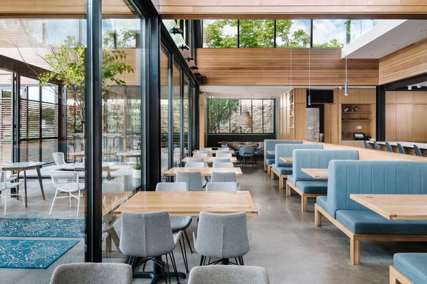 A spacious contemporary commercial dining and lounge interior with extensive floor-to-ceiling glazing, exposed timber ceiling and wall lining, polished concrete flooring, and abundant natural light. The room contains pale blue upholstered booth seating, timber tables, grey dining chairs, and a separate glazed dining area with patterned rugs and outdoor greenery visible beyond.