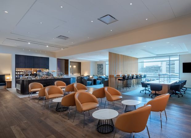 A spacious contemporary commercial hospitality and lounge interior with a polished bar, dark timber cabinetry, marble-look counter, clusters of tan leather lounge chairs, small round tables, blue sofas, and a long communal high table. Floor-to-ceiling glazing opens the bright rear dining/work area to an urban view. A slatted timber screen subtly separates the zones, while recessed downlights and linear ceiling lighting provide even illumination. The space has warm timber flooring, neutral walls, and a refined business-lounge atmosphere.