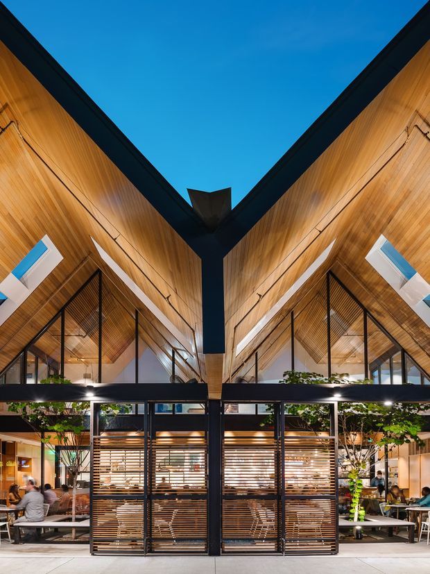 A dramatic symmetrical interior of a contemporary commercial space, viewed toward a central dark steel structural junction beneath a high V-shaped timber-lined ceiling. Full-height glazing, black metal framing, timber louvres, built-in seating, indoor trees, and tables suggest a restaurant, café, or hospitality venue. The exposed pitched roof form and warm timber interior are strongly emphasized.