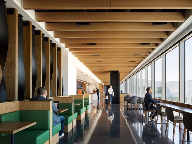 A contemporary café or hospitality interior with a long, linear layout. Green upholstered booth seating and timber tables occupy the left side, while a row of loose dining tables and chairs lines the full-height glazed facade on the right. The space features a highly articulated exposed timber ceiling with regularly spaced beams, dark polished flooring, vertical timber and dark acoustic wall elements, pendant lighting, and a service counter in the distance. People are seated and walking through the space, with bright views visible beyond the glazing.