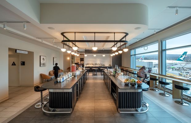 A contemporary airport lounge or hospitality space viewed from the main seating and service area. Two long, dark island-style counters with ribbed bases run toward a rear bar and preparation zone, topped with drinks, snacks, and tableware. Warm globe pendant lighting hangs within a black rectangular frame beneath a large curved ceiling bulkhead. Floor-to-ceiling glazing on the right overlooks an airport apron and aircraft. The interior uses a restrained palette of grey, beige, black, and warm timber tones, with bar stools and lounge seating along the window.