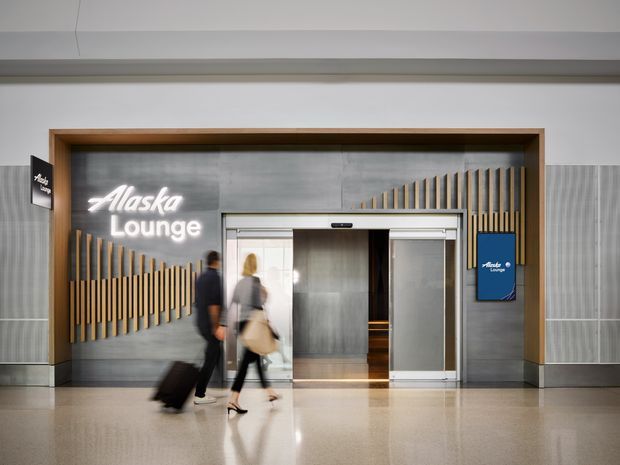 A polished airport-style interior entrance for an Alaska Lounge, featuring a central elevator or doorway opening framed in warm timber, grey metallic wall panels, decorative vertical timber slats, illuminated white signage, and a reflective light floor. Blurred travelers with luggage pass in front of the entrance.
