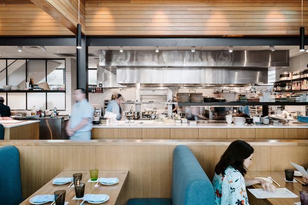 A bright contemporary restaurant interior viewed from the dining area toward an open commercial kitchen. The space features a long timber-fronted service counter, stainless-steel commercial cooking and extraction equipment, open shelving with dishes and supplies, turquoise upholstered booth seating, and timber-clad ceiling and upper wall surfaces. Diners and staff are present, with large glazed partitions visible at the left.