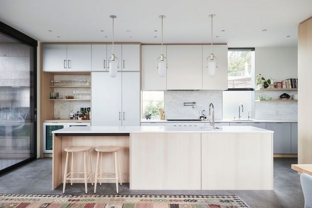 Bright contemporary kitchen with a large pale timber island, integrated cabinetry, white benchtops, marble-look splashback, pendant lighting and three timber bar stools. Full-height glazing and a window bring in natural light, while open shelving and a patterned rug add warmth and texture.