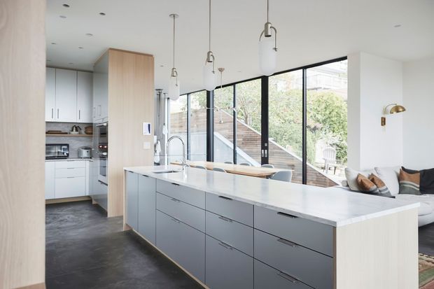 Bright open-plan kitchen with a large central island, pale timber end panels, matte grey cabinetry, and a white stone benchtop. A wall of black-framed glazing opens the kitchen toward an outdoor area, while pendant lights hang above the island and a lounge seating area is visible to the right.