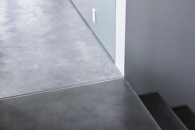 Close-up architectural view of a minimalist stair landing and staircase. A large grey concrete- or stone-like landing transitions to dark perforated metal stair treads, bordered by smooth light and dark grey walls with a crisp white vertical trim.