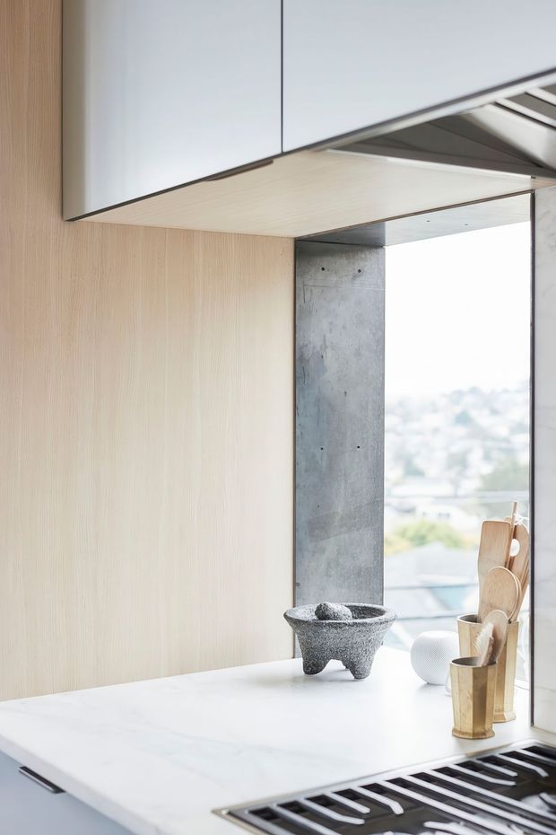 Close-up of a refined contemporary kitchen interior, focused on a pale stone kitchen island and integrated black cooktop beneath sleek white overhead cabinetry. Light timber wall panelling, a raw concrete structural column, and a large bright window create a clean, minimalist composition. Small decorative objects sit on the benchtop, with a softly blurred exterior view beyond the glazing.
