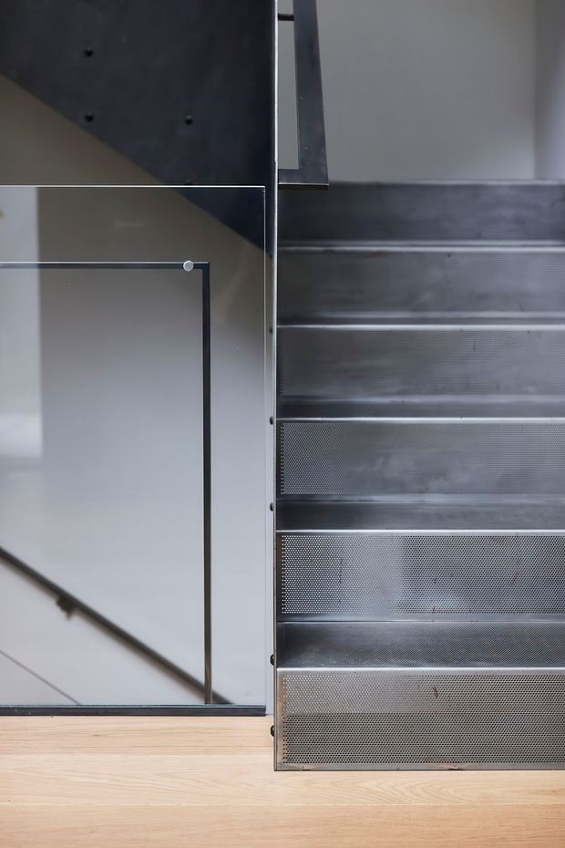 Close-up architectural detail of a contemporary stairway with perforated dark metal stair treads and risers beside a clear glass balustrade with slim dark framing. Pale timber flooring and a minimalist light-colored wall create a restrained, industrial-modern composition.