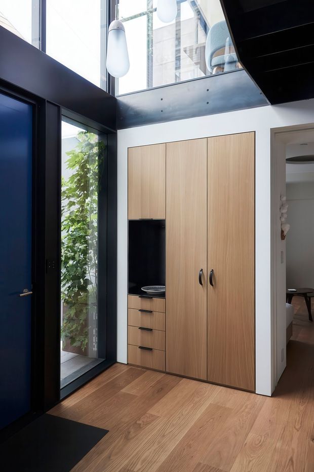A bright contemporary entryway with a dramatic double-height void. The space features a dark blue-black entry door and surrounding architectural framing, floor-to-ceiling glazing, a bridge or upper-level opening with a dark metal soffit, pendant lights, and extensive light timber built-in cabinetry with integrated drawers and a recessed black display niche. Timber flooring continues toward an adjacent room visible through the doorway.