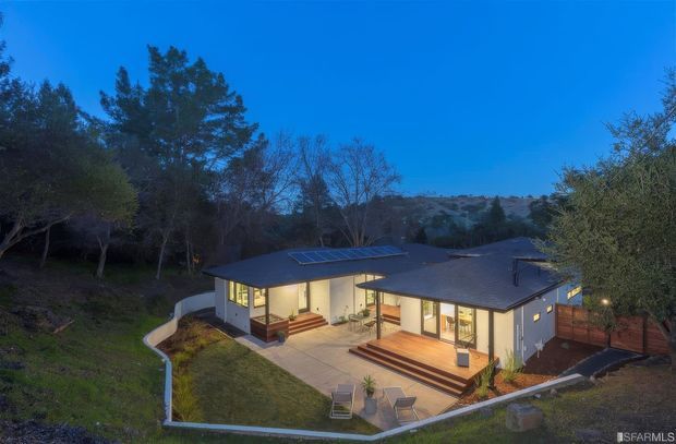 Aerial dusk view of a low-profile single-storey contemporary home set into a wooded, sloping site. The dark, gently pitched roof includes a visible solar-panel array, while broad glazed openings face a paved central terrace with timber steps, outdoor seating, and landscaped garden edges. Warm interior lighting contrasts with the cool blue evening sky and surrounding trees.