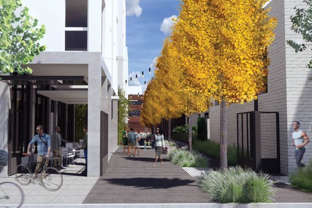 A landscaped pedestrian courtyard or laneway between contemporary multi-storey buildings. The scene features white and grey masonry facades, large glazed openings, a covered outdoor seating area with bicycles, dark paved pedestrian surfaces, ornamental grasses, mature golden deciduous trees, and suspended string lighting. People are walking and sitting within the shared outdoor space.