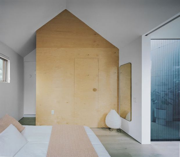 A contemporary bedroom with a minimalist built-in plywood feature wall shaped like a house gable, incorporating a concealed door. The room has a low bed with white bedding and a pale blush runner, light timber flooring, white walls, a sculptural floor lamp, and a tall rounded mirror. A ribbed translucent glazed opening is visible at right, with a small window at left.