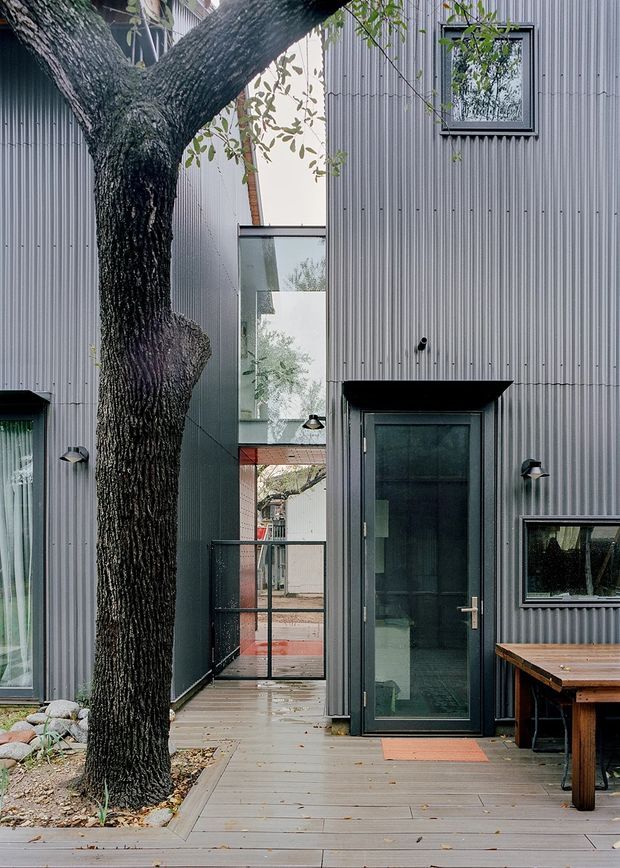 A modern multi-level residential facade viewed from a timber deck and entry walkway. The building is clad in vertically ribbed grey metal, with dark-framed glazed doors and windows, exterior wall lights, and a narrow glazed link or courtyard passage. A large established tree grows through the deck beside a small rock garden, while a timber outdoor table occupies the right foreground. The composition emphasises an urban courtyard relationship between mature landscape and contemporary architecture.