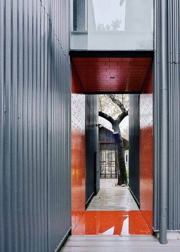 A narrow covered exterior passage between corrugated metal-clad walls, with vivid red glazed brick or tile lining the side walls, soffit, and lower floor threshold. A timber walkway leads toward a courtyard-like opening containing a mature tree and glazed architectural elements. The composition highlights a dramatic framed transition, reflective wet surfaces, and strong contrast between charcoal grey metal and red ceramic surfaces.
