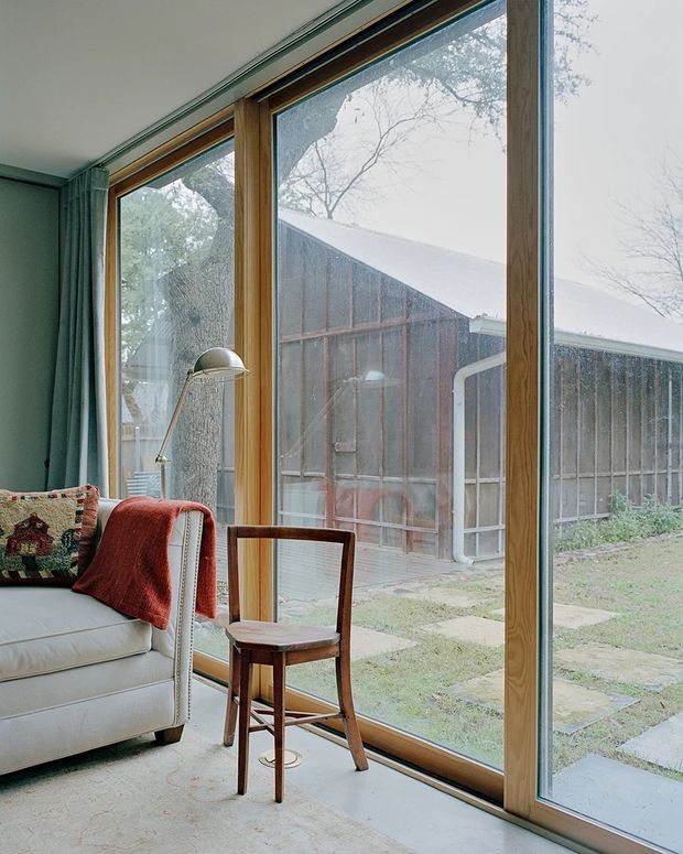 A calm living room corner with full-height timber-framed glazing overlooking a rustic outbuilding and garden. A light upholstered sofa with a patterned cushion and rust-red throw sits beside a slim floor lamp, while a small vintage wooden chair is positioned near the window. The room has a pale carpet, muted green walls and curtains, and strong natural light filtered through the glazing.