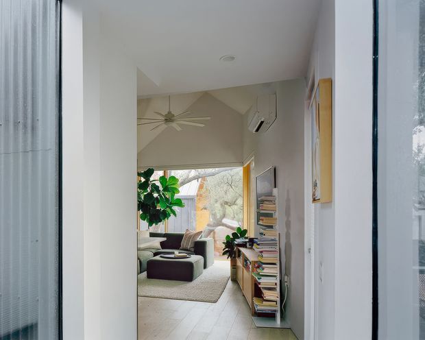 View from a bright hallway or entry toward a compact living room. The living area has pale walls, light timber flooring, a large window with roller blind, a ceiling fan, low contemporary seating, a neutral rug, abundant indoor plants, and an open shelving unit filled with books. The image also shows a simple white interior shell with a high, angled ceiling transition and glimpses of exterior glazing.