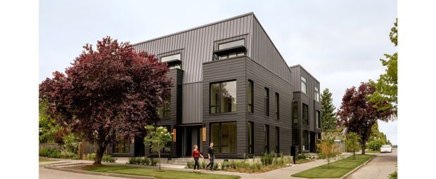 Exterior view of a contemporary multi-storey residential building with dark vertical cladding, projecting glazed bays, and a steeply pitched gable roof. The building sits along a landscaped suburban street with mature ornamental trees and pedestrian paths.