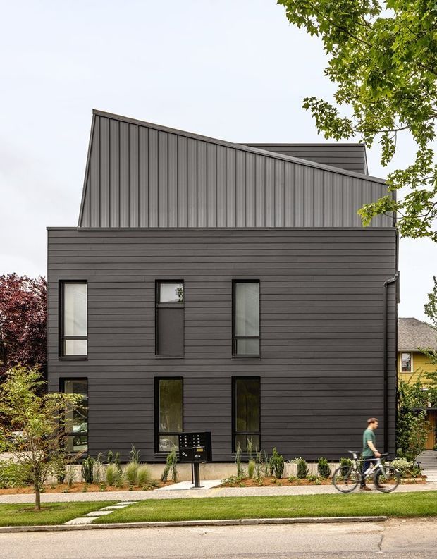 Front exterior view of a contemporary multi-storey residence with a dark charcoal horizontal-clad lower facade, vertically ribbed metal upper volume, sharply angled roofline, and tall narrow windows. A landscaped verge, pedestrian path, and street frontage sit in front of the house, with mature trees framing the scene.