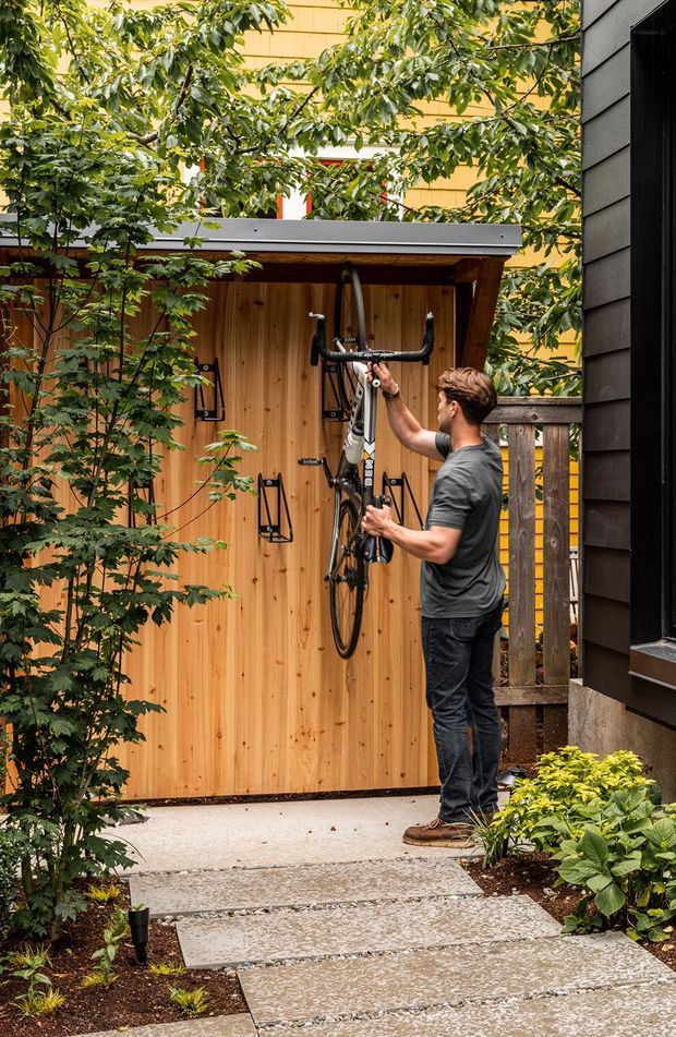 A landscaped residential side path leads to a compact timber bicycle storage shed. The shed has vertical natural-wood cladding, a shallow dark metal roof, and open double doors with wall-mounted bicycle racks. A person is placing a road bicycle inside. Dense planting surrounds the concrete stepping-stone walkway, while a dark-clad building wall and timber fence frame the scene.
