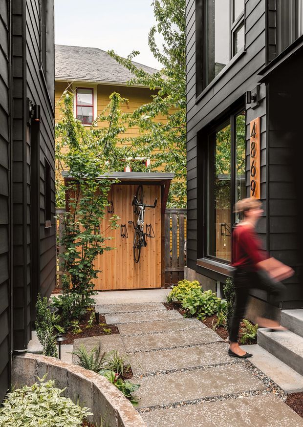 A narrow landscaped urban courtyard walkway between two dark-clad residential buildings. Large rectangular concrete pavers form the path, bordered by planted garden beds and low retaining walls. A timber bicycle storage shed stands at the rear, with a glimpse of a yellow-clad neighbouring building beyond. The contemporary black horizontal cladding, large windows, entry steps, and vertical address plaque define the architecture; a blurred person is walking through the foreground.