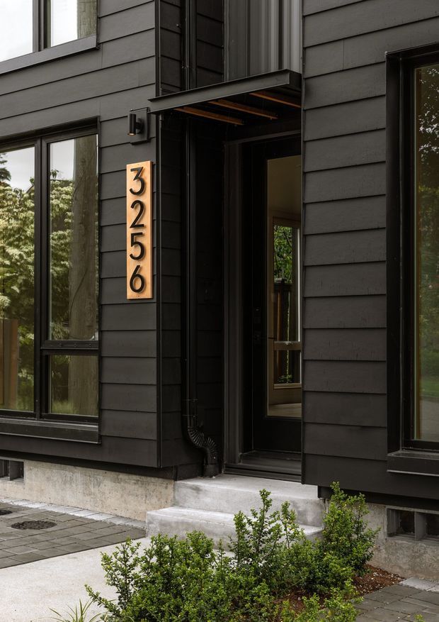 Close-up exterior view of a contemporary building entrance with dark horizontal cladding, black-framed glazing, a recessed glazed front door, concrete steps and foundation, and a vertical timber-toned address plaque reading 3256. Low shrubs and paved paths frame the entry.