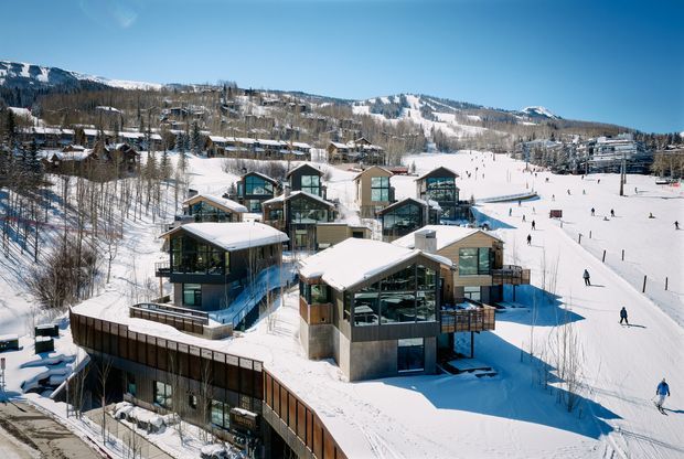 Aerial view of a contemporary alpine resort development set directly beside snow-covered ski slopes. Multiple modern chalet-style buildings feature steeply pitched roofs, extensive dark-framed glazing, timber-toned cladding, and projecting balconies, with skiers visible across the surrounding mountain landscape.