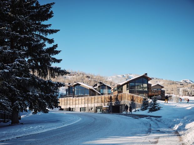 Snow-covered mountain resort buildings arranged along a gently curving access road, with dark timber-and-glass facades, steep gabled roofs, large glazed openings, and surrounding evergreen trees. A snowy alpine landscape and ski activity are visible in the background under a clear blue sky.
