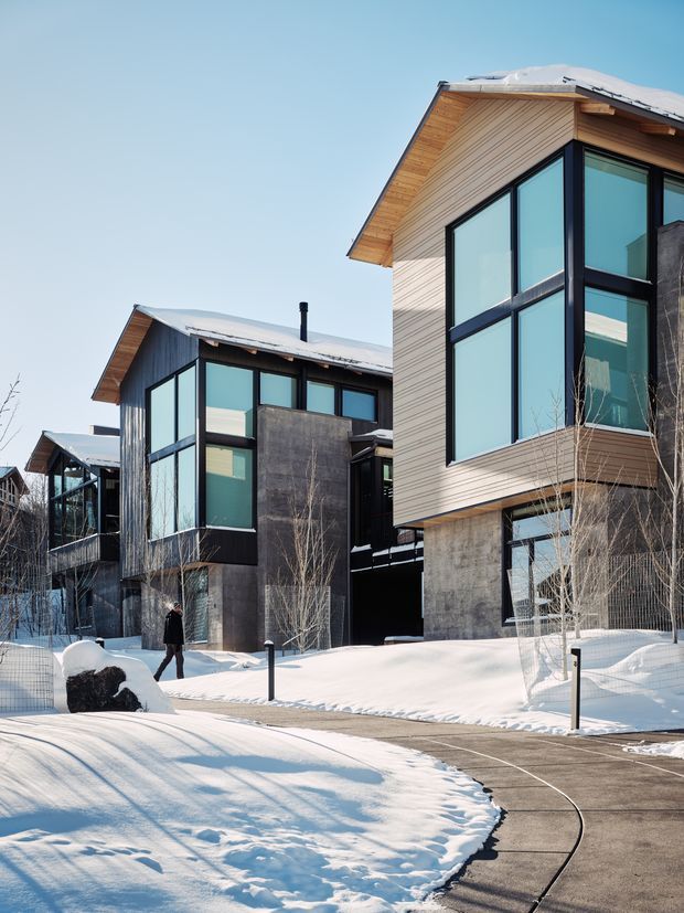 Exterior view of a contemporary multi-storey residential development in a snowy setting. The buildings feature steeply pitched gable roofs with standing-seam metal roofing, expansive black-framed glazing, timber-clad upper levels, and exposed concrete lower sections. A paved pedestrian path and landscaped snow-covered grounds run between the dwellings.
