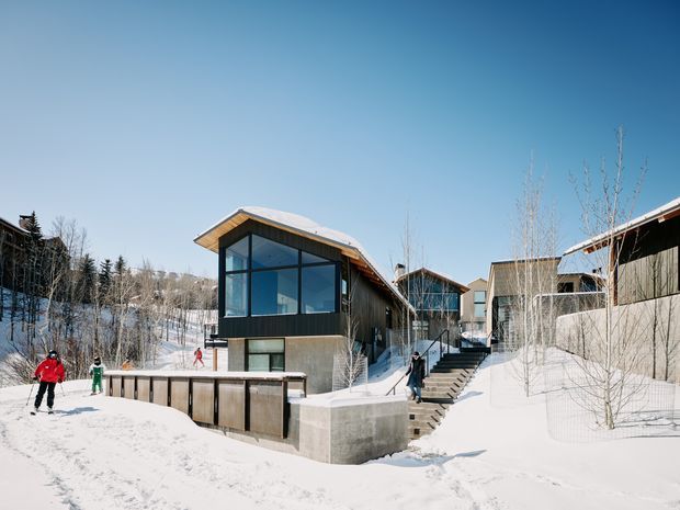 Exterior view of a contemporary multi-building development in a snowy, wooded setting. The buildings feature dark vertical cladding, extensive black-framed glazing, angular pitched roof forms with timber soffits, concrete retaining walls, and connected pedestrian stairs and walkways. People are skiing or walking through the snow-covered grounds.