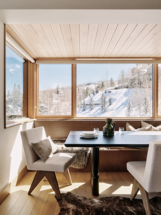 A bright, compact dining nook with a dark rectangular dining table and upholstered timber dining chairs, enclosed by large timber-framed windows overlooking a snowy alpine landscape. The room features warm timber wall and ceiling panelling, timber flooring, neutral upholstery, and a fur rug, creating a refined mountain-lodge atmosphere.