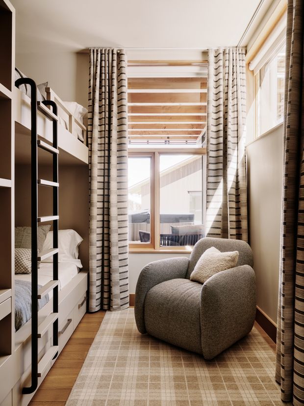 A compact, light-filled bunk bedroom with built-in white bunk beds and black ladder shelving along the left wall. A sculptural textured taupe armchair sits on a large neutral plaid rug beside full-height patterned curtains. Warm timber flooring, timber-framed glazing, and exposed timber ceiling battens create a cosy contemporary cabin-like atmosphere.