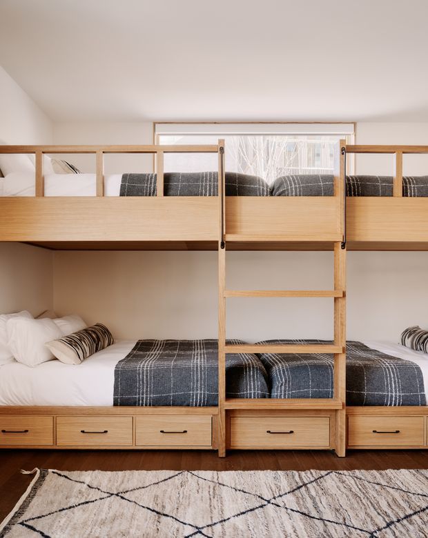 A compact bunk-bedroom with a custom built-in timber bunk bed spanning the wall. Two lower and two upper sleeping berths have white bedding with dark grey plaid blankets and striped cushions. Integrated drawers run beneath the lower bunks, and a central timber ladder connects the levels. A large geometric neutral rug covers the dark timber floor, while warm white walls and a rear window create a bright, cozy atmosphere.