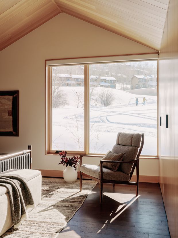 A warm, sunlit living room with a steep vaulted ceiling lined in pale timber boards, cream walls, and a large picture window overlooking a snowy residential landscape. The room contains a mid-century-style upholstered lounge chair, a light upholstered sofa or ottoman, a patterned rug, a radiator, a vase with branches, and built-in white cabinetry. Dark timber flooring contrasts with the light interior and winter view.