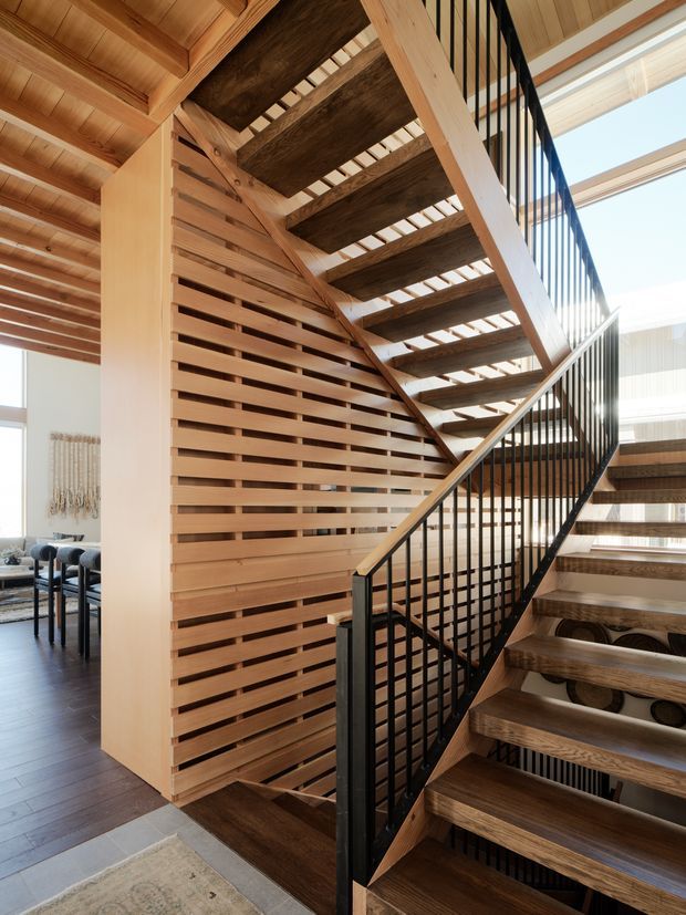 Interior view of a contemporary timber staircase with open-riser wooden treads, black metal balustrades, and a slatted timber feature wall. The stair rises through a bright, high-ceilinged space with exposed timber structure and glimpses of a dining area beyond.