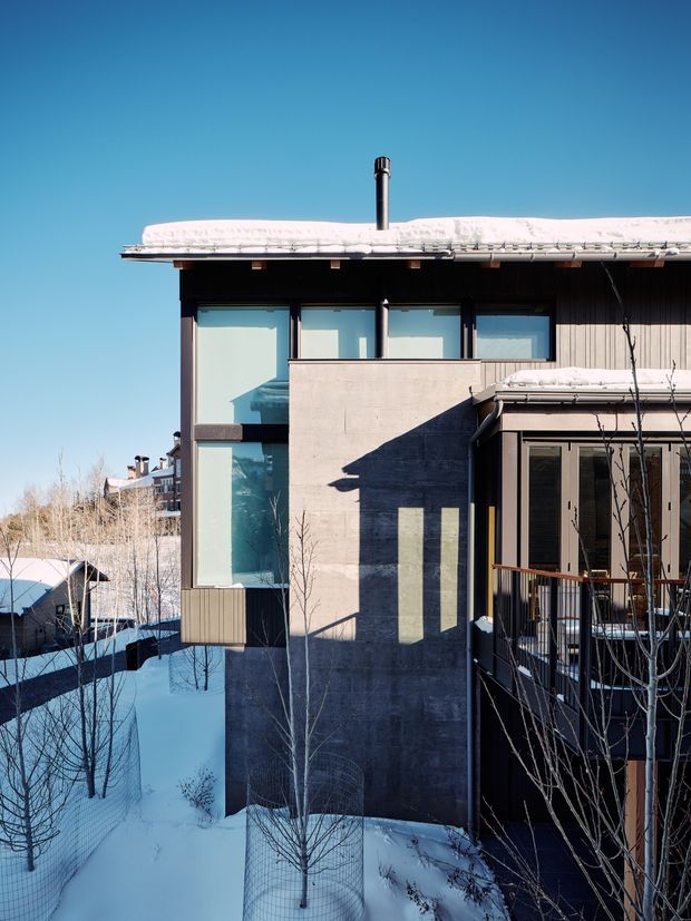 Exterior view of a contemporary multi-level house in a snowy setting. The building combines dark vertical cladding, exposed concrete walls, extensive floor-to-ceiling glazing, and a low-pitched roof covered in snow. A chimney, projecting balcony, timber-toned window and door frames, and a partially enclosed outdoor deck are visible. Bare deciduous trees and deep snow surround the home.